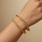 Two gold rope-style bangle bracelets worn on a person's wrist against a warm beige background.