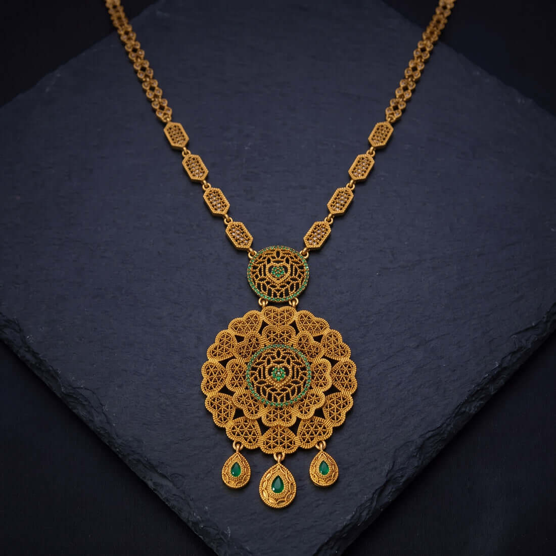 An ornate Antique Gold Big Floral Heart Pendant Long Necklace with a large filigree pendant and three teardrop charms, accented with small green stones, displayed on a dark slate surface.