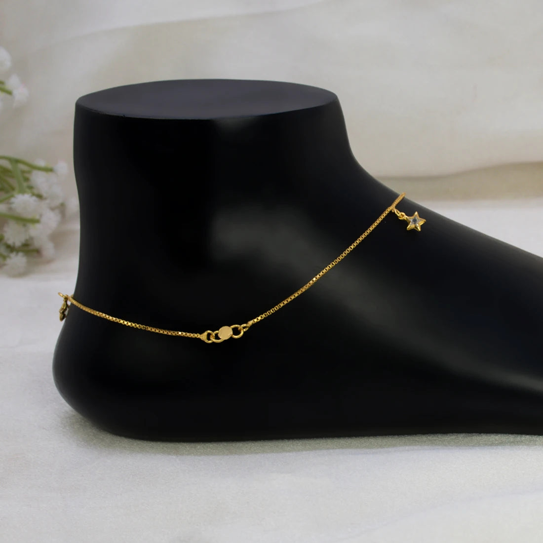 A delicate gold-plated crystal star charm anklet with a small star charm and a heart-shaped connector, displayed on a black foot-shaped jewelry stand against a soft white fabric background.