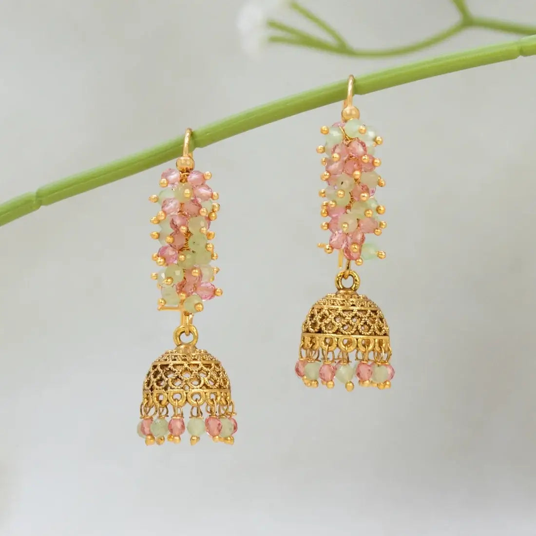 A pair of gold-toned traditional Indian Antique Gold Plated Colourful Beaded Jhumka with pink and light green stone clusters, hanging from a green stem against a soft white background.