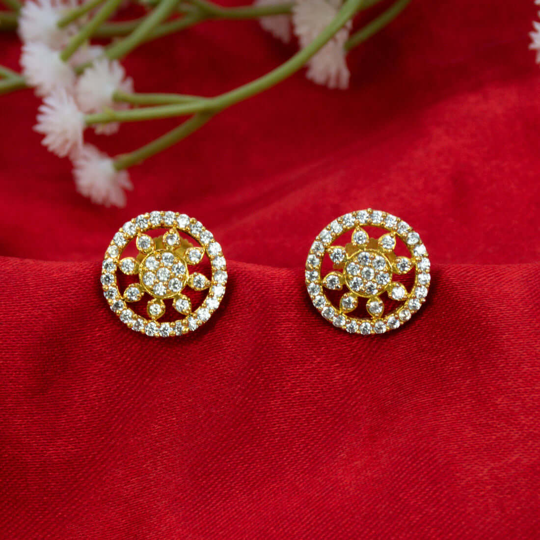 A pair of Gold-Plated CZ Sunburst Stud Earrings with circular floral designs set with clear stones, displayed on a textured red fabric background with small white flowers in the upper corner.
