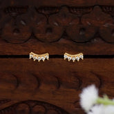A pair of Gold-Plated Cubic Zirconia Arc Second Stud Earrings with small clear stones, displayed on a dark carved wooden surface.