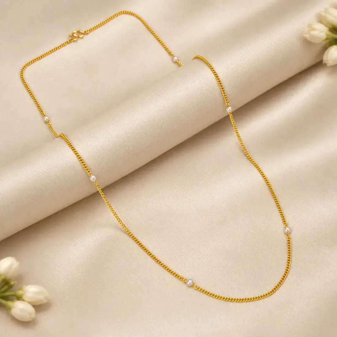 A delicate Gold-Plated Curb Link White Pearl Chain with small white pearl beads spaced along the chain, displayed on a draped beige satin fabric with white flowers in the corners.