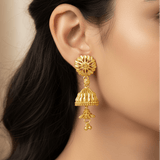 A close-up of a woman's ear wearing a detailed Gold-Plated Two-Step Floral Jhumka Earrings with a floral top, bell-shaped dangle, and small hanging bells.