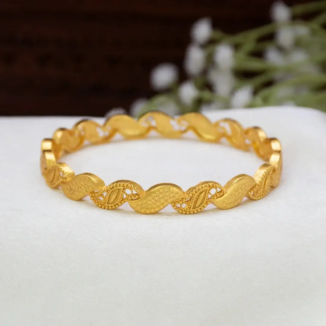 A Gold-Plated Wave Leaf Designer Bangle For Women with a textured, wavy interlocking link design, displayed on a white surface with a soft-focus background.