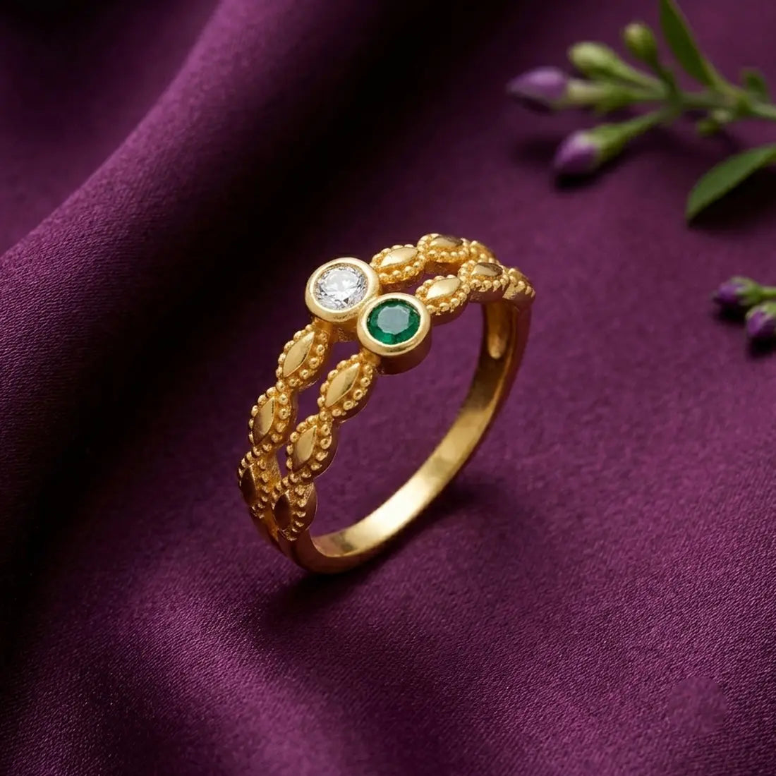 A gold ring with an intricate leaf-patterned band, featuring a clear round gemstone and a green oval gemstone, displayed on a draped purple fabric background.