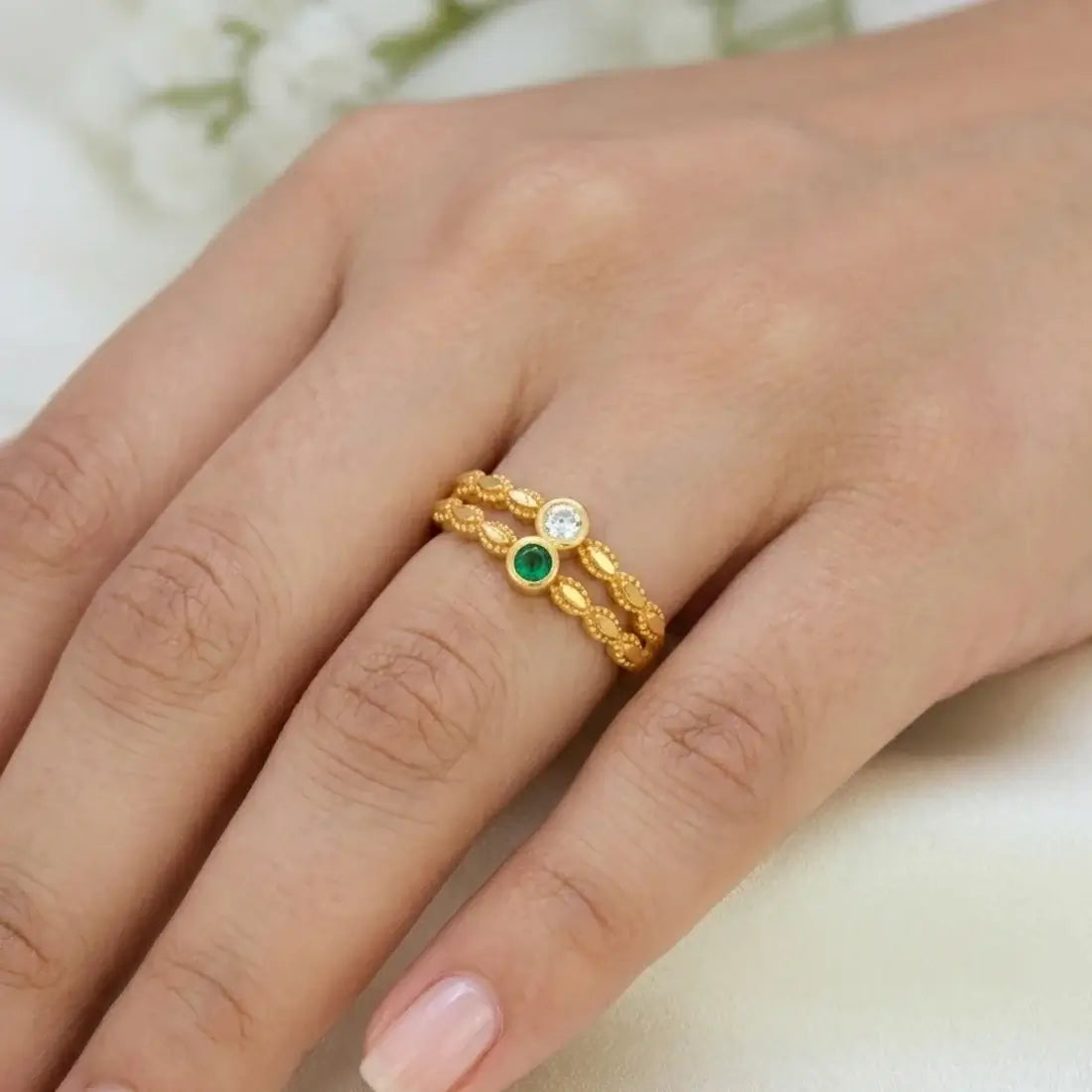A close-up of a hand wearing a delicate gold ring with three small circular stones, including one green stone and one clear stone, set against a soft light background.