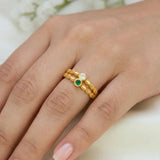 A close-up of a hand wearing a delicate gold ring with three small circular stones, including one green stone and one clear stone, set against a soft light background.