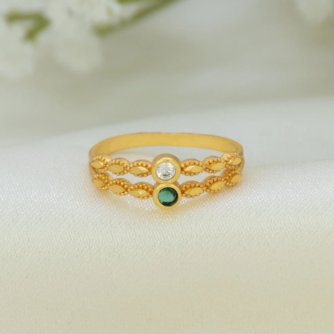 A gold-plated ring with a textured, braided filigree design, featuring a clear round-cut stone on one side and a small green oval stone on the other, resting on a soft white fabric background.