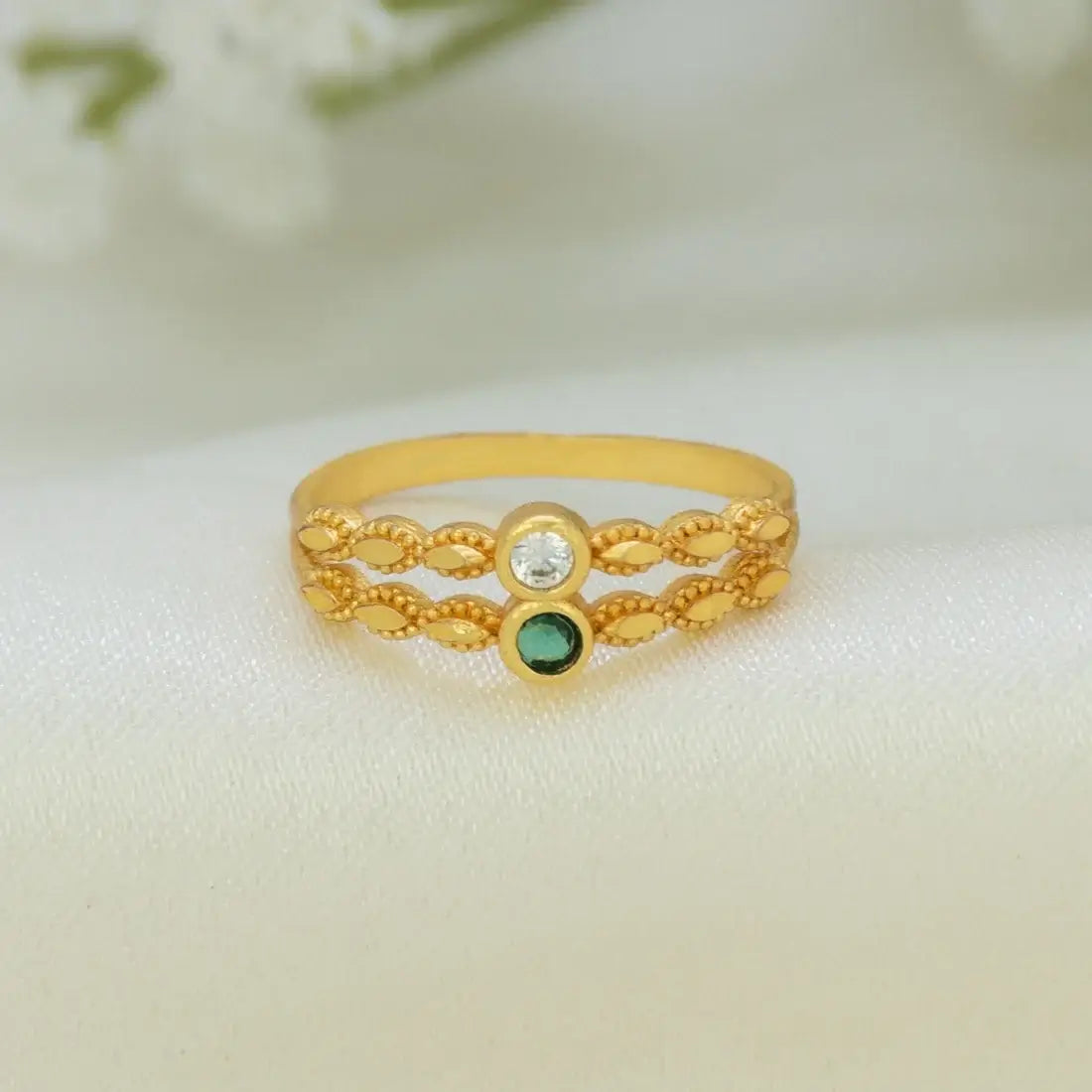 A gold-plated ring with a textured, braided filigree design, featuring a clear round-cut stone on one side and a small green oval stone on the other, resting on a soft white fabric background.