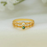 A gold-plated ring with a textured, braided filigree design, featuring a clear round-cut stone on one side and a small green oval stone on the other, resting on a soft white fabric background.