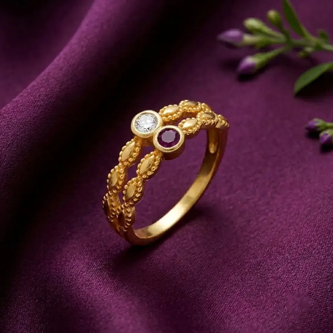 A gold ring with intricate leaf-like detailing, featuring a clear round gemstone and a round purple gemstone, displayed on a draped purple fabric background.