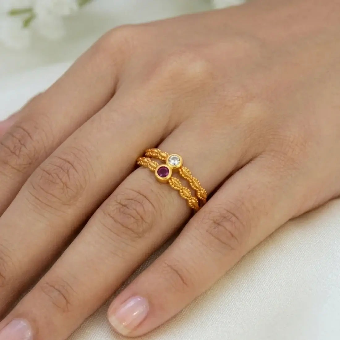 A close-up of a hand wearing a delicate gold ring with two twisted bands and small round gemstones, one clear and one purple, on a soft light background.