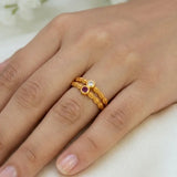 A close-up of a hand wearing a delicate gold ring with two twisted bands and small round gemstones, one clear and one purple, on a soft light background.