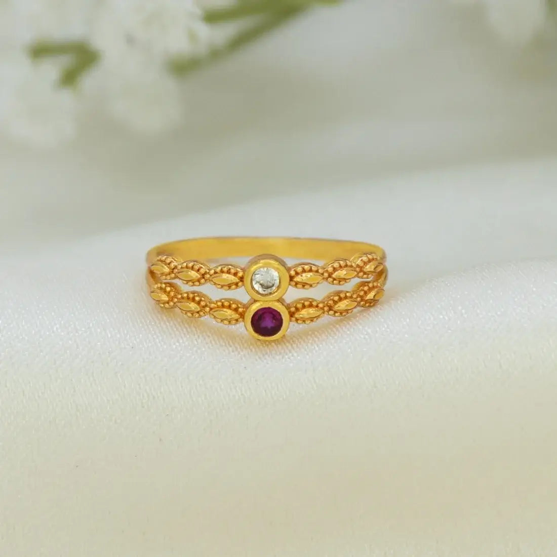 A gold ring with a textured, twisted band design, featuring a top setting with a clear round gemstone and a bottom setting with a round purple gemstone, displayed on a soft white fabric background.