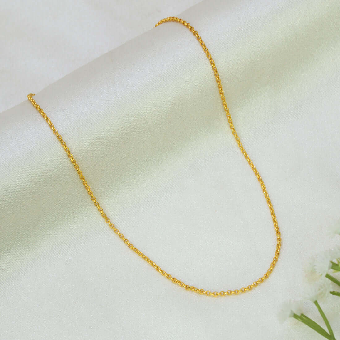 A Gold Plated Rolo Baby Chain for Kids & Petite Adults draped in a U-shape on a soft, light cream-colored fabric background with a small white flower in the corner.