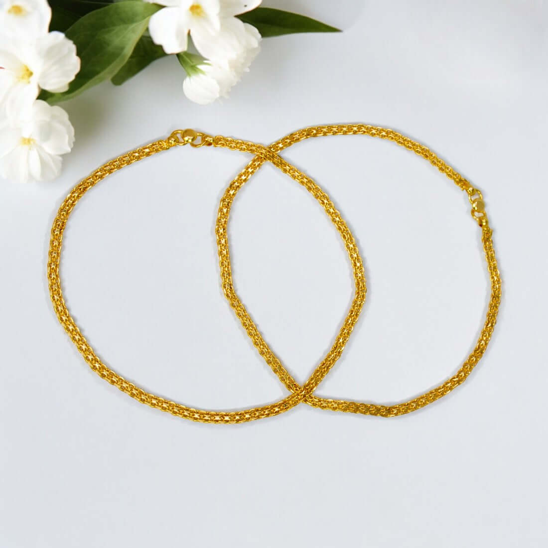 Trendy Gold Plated Urvashi Chain Anklets on a light gray background with white flowers and green leaves.