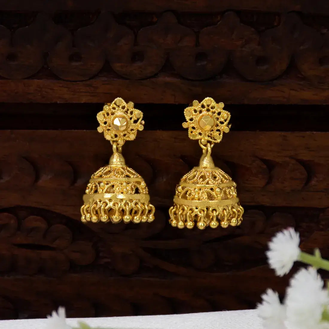 A pair of ornate Grand Traditional Gold-Plated Filigree Jhumka Earrings displayed against a dark carved wooden background with small white flowers in the foreground.