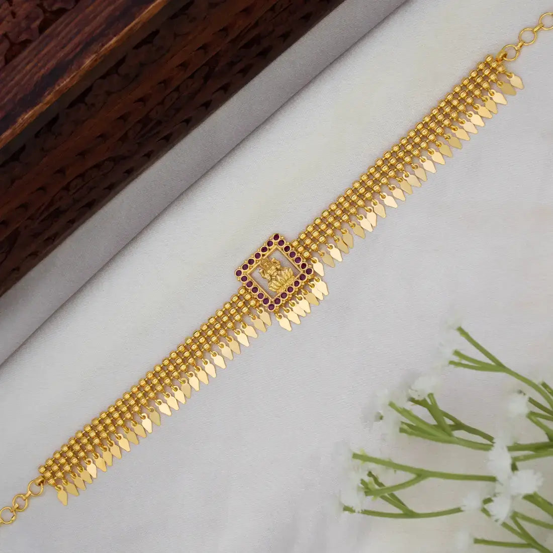A Beaded Gold Plated Ruby Square Lakshmi Elakkathali Choker, displayed diagonally on a white textured surface with a dark wooden block and small white flowers as props.