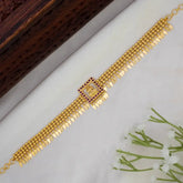 A Beaded Gold Plated Ruby Square Lakshmi Elakkathali Choker, displayed diagonally on a white textured surface with a dark wooden block and small white flowers as props.