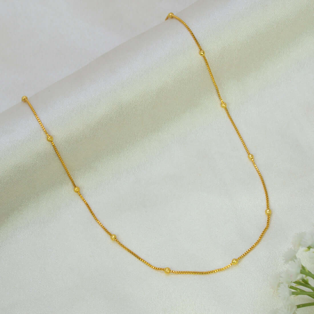 A delicate short gold-plated beaded box chain - 15 Inches with small spaced bead accents, displayed on a soft white fabric background.