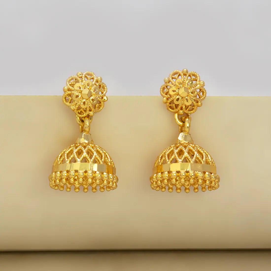 Pair of gold-plated jhumka earrings on a beige surface with a light gray background
