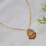 A gold chain necklace with a decorative pendant featuring a gold-toned Lakshmi central motif and ruby stones, displayed on a soft white fabric surface.