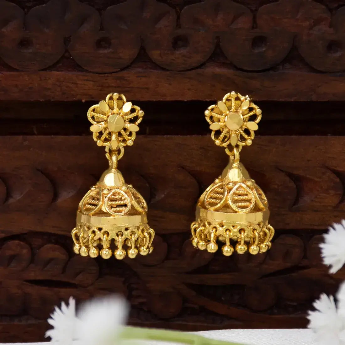 A pair of South Indian Medium Jhumki Earrings For Women with floral tops and bell-shaped dangling bells, displayed on a carved dark wood surface.