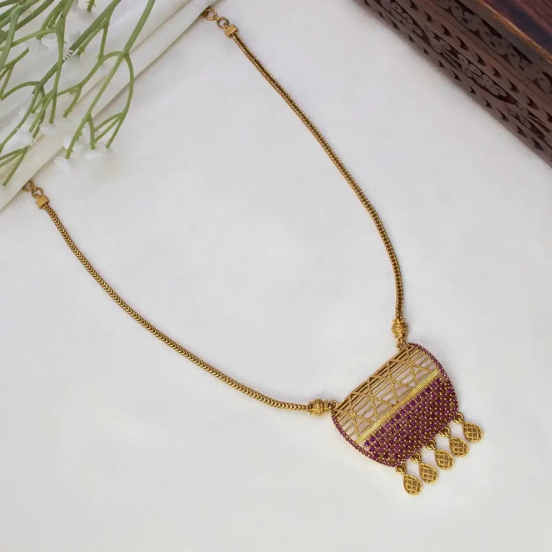 Victorian Style Antique Stone Mesh Pendant Necklace with a semi-circular cutout design, accented with deep pink stones and dangling teardrop charms, displayed on a white surface with decorative greenery.