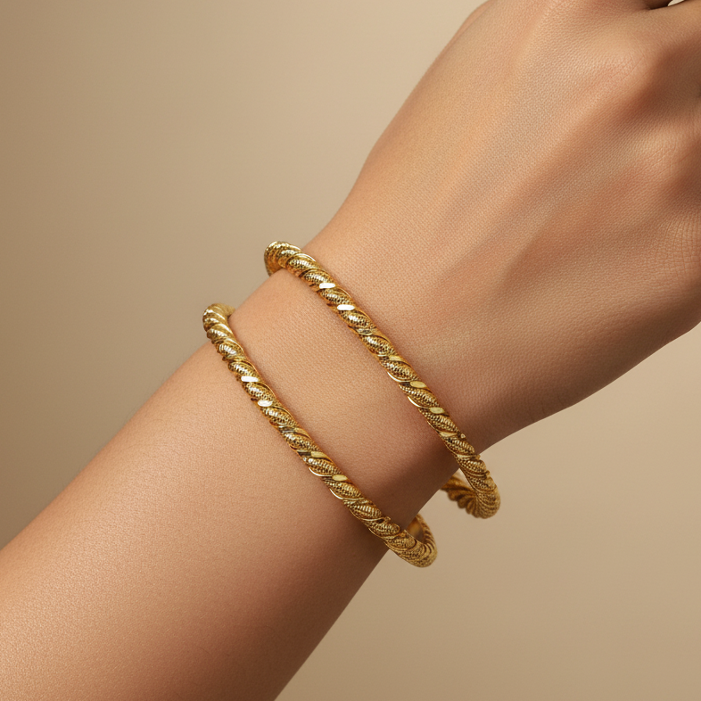 Two gold rope-style bangle bracelets worn on a person's wrist against a warm beige background.