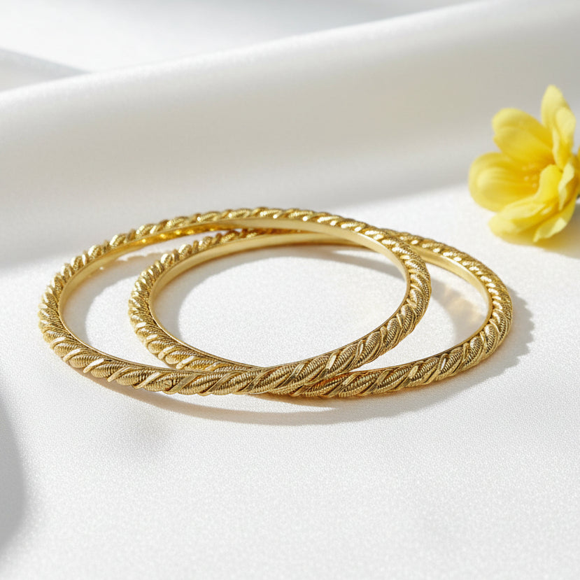 Two gold-colored twisted rope bangles with a textured, braided pattern, displayed overlapping on a reflective white surface.