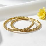Two gold-colored twisted rope bangles with a textured, braided pattern, displayed overlapping on a reflective white surface.