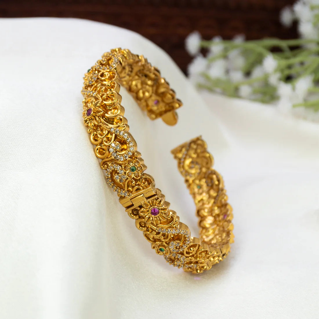 An Antique Floral Multi-colour Stone Cuff Bangle with small inset stones and tiny colored gem accents, displayed on a white fabric surface with blurred white flowers in the background.