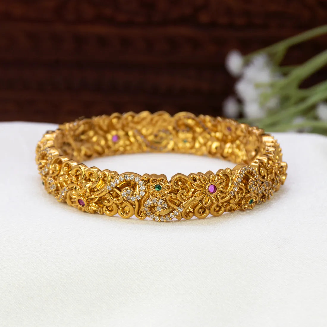 An Antique Floral Multi-colour Stone Cuff Bangle with intricate filigree patterns, set with small pink, green, and clear stones, resting on a white textured surface.