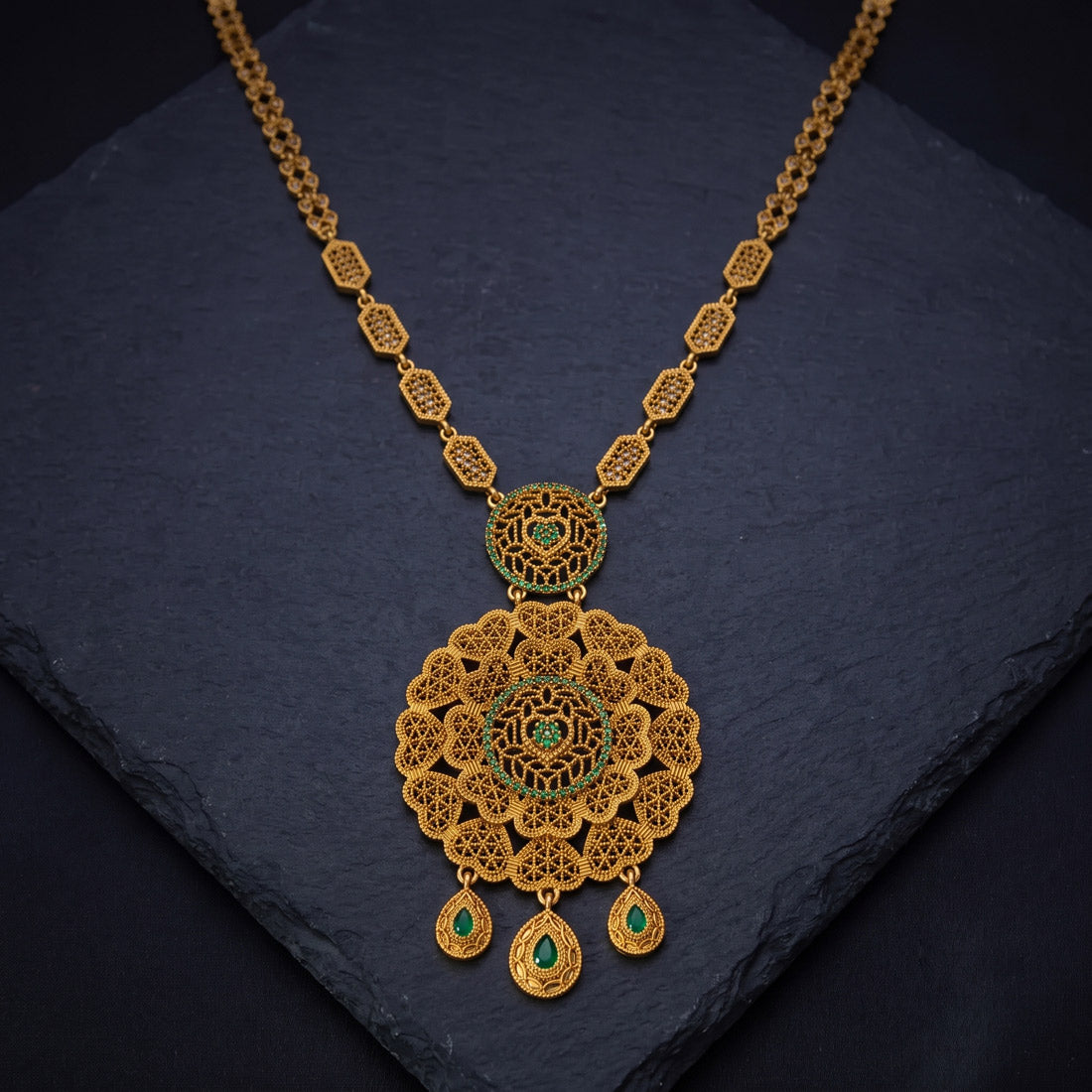 An ornate Antique Gold Big Floral Heart Pendant Long Necklace with a large filigree pendant and three teardrop charms, accented with small green stones, displayed on a dark slate surface.