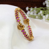 An Antique Gold Plated Ruby Stone Peacock Bangle adorned with oval ruby stones and a decorative peacock charm, displayed on a white fabric surface with blurred white flowers in the background.
