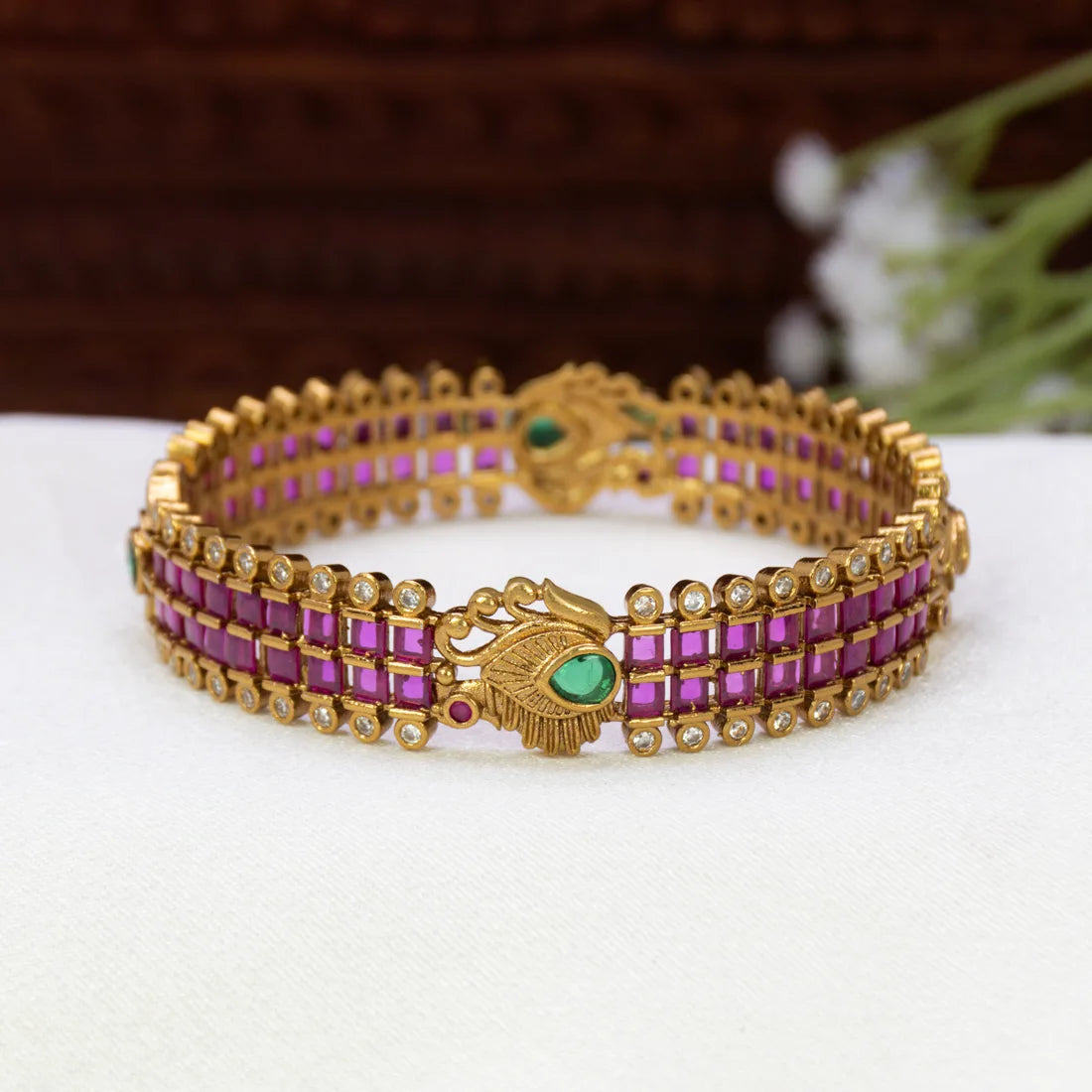 Antique Peacock Kemp and CZ Stone Bangle featuring rows of rectangular ruby red stones and small clear stones, with a central decorative motif and a green stone accent, displayed on a white surface.