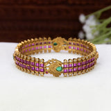 Antique Peacock Kemp and CZ Stone Bangle featuring rows of rectangular ruby red stones and small clear stones, with a central decorative motif and a green stone accent, displayed on a white surface.