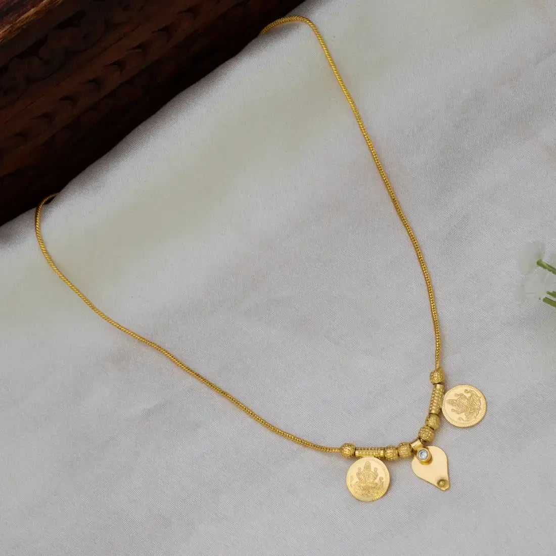 A delicate gold plated chain necklace with two coin-style pendants and a small leaf charm, displayed on a soft white fabric surface.