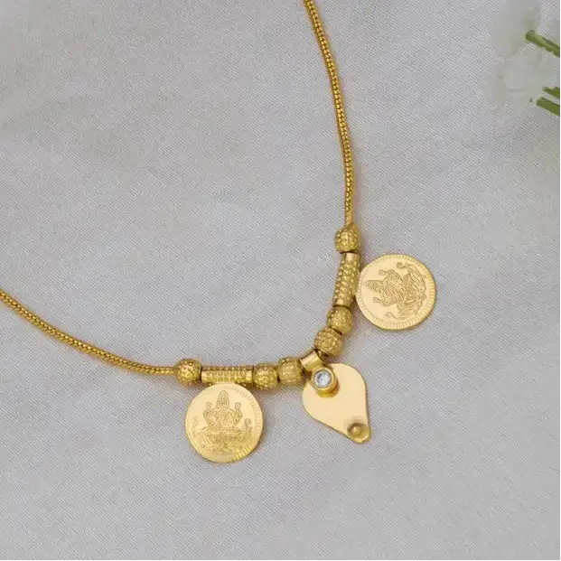 A delicate gold chain necklace with two coin-style pendants and a small thali charm, displayed on a soft white fabric surface.