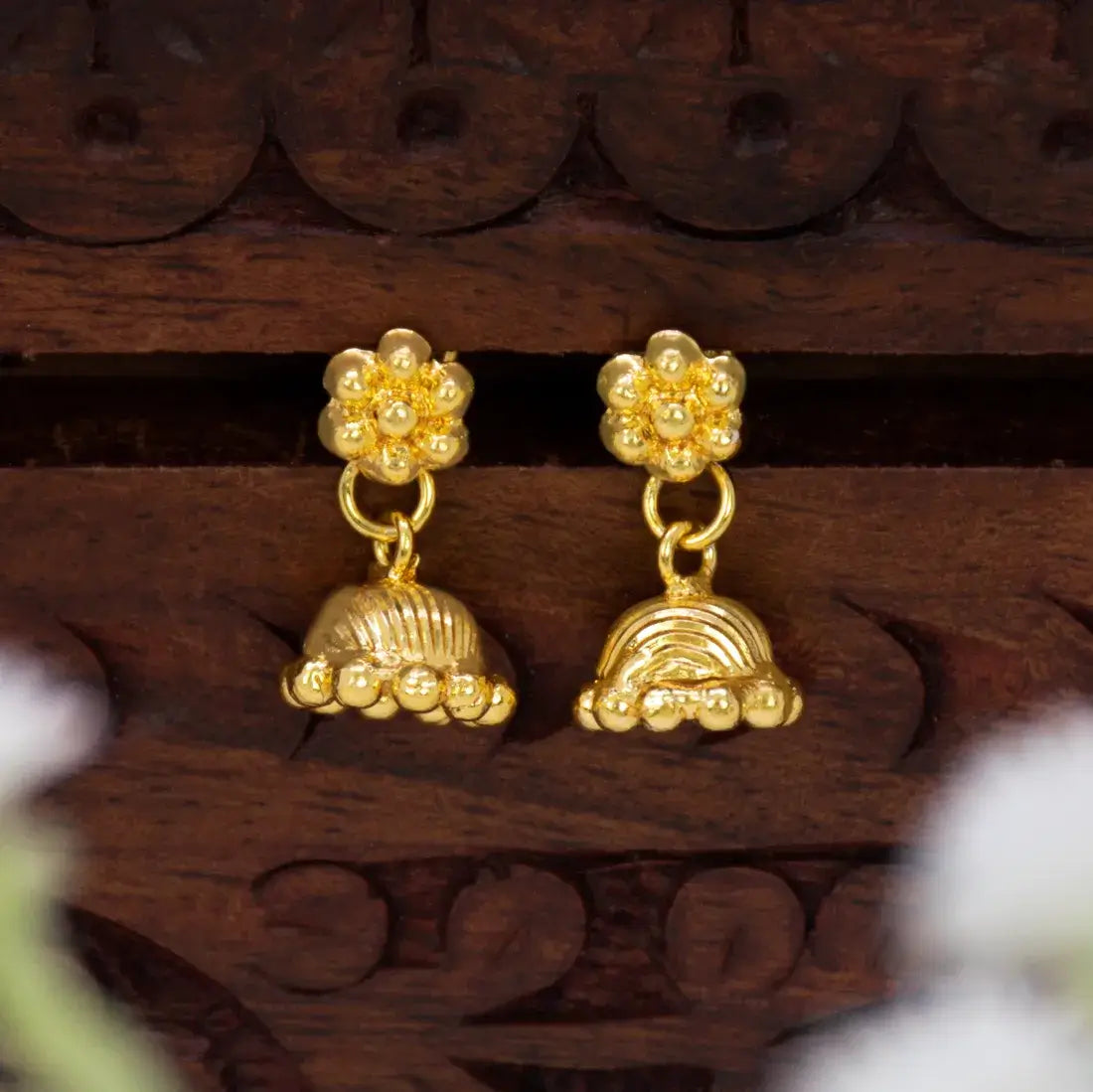 A pair of Cute Gold-Plated Kids Floral Jhumka Earrings with floral studs and bell-shaped dangling charms, displayed on a carved dark brown wooden surface.