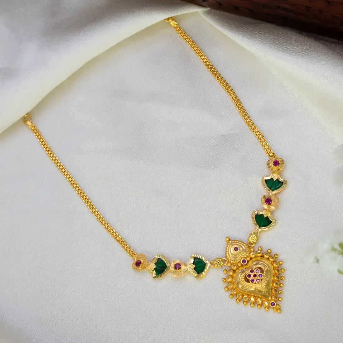 Gold-Plated Palakka Heart Pendant Necklace with a large ornate pendant, green Palakka stone, and small ruby stones, displayed on a soft white fabric background.
