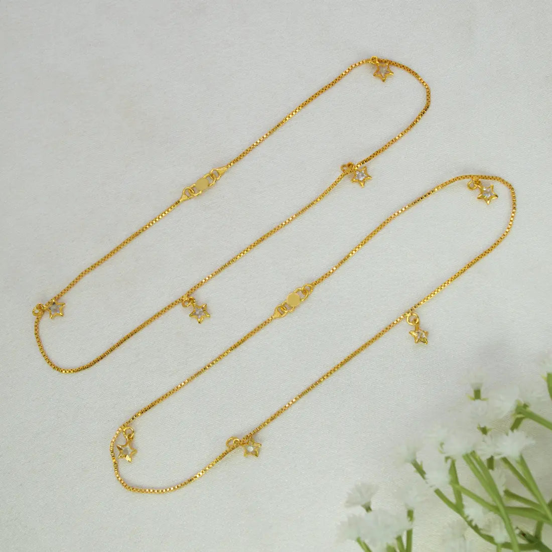 Delicate Gold-Plated Crystal Star Charm Anklets with small bow and star charms, arranged diagonally on a light textured background with white flowers in the corner.