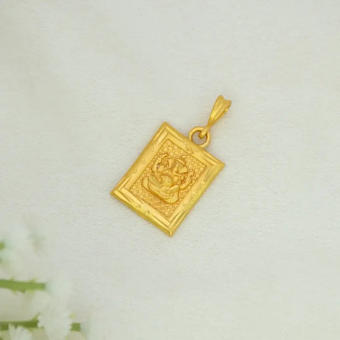 Devotional Gold Plated Rectangle Ganesha Pendant with an embossed elephant design, shown on a light textured background with a soft white flower prop in the lower left corner.