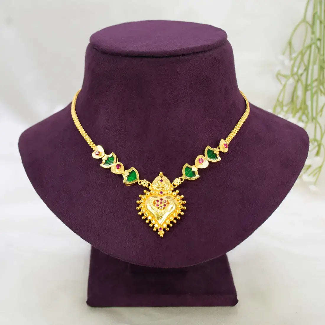 Gold-Plated Palakka Heart Pendant Necklace with a heart-shaped pendant and green Palakka and ruby stones, displayed on a purple velvet jewelry bust against a light background.