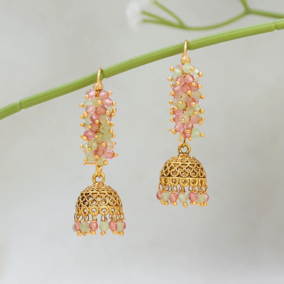 A pair of gold-toned traditional Indian Antique Gold Plated Colourful Beaded Jhumka with pink and light green stone clusters, hanging from a green stem against a soft white background.