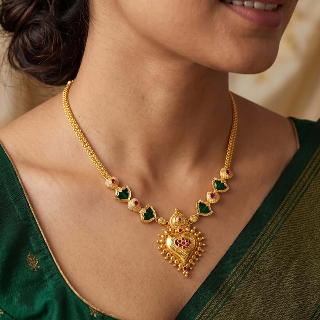 A close-up of a woman wearing a Gold-Plated Palakka Heart Pendant Necklace with a heart-shaped pendant and green palakka and Ruby stones, paired with a dark green traditional garment.