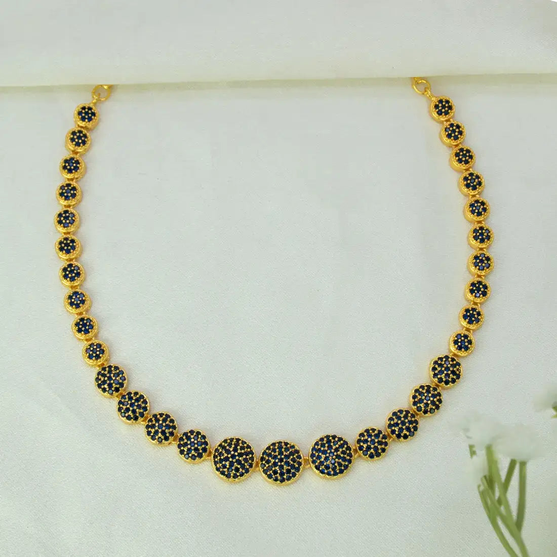 A Gold-Plated Round Design Stone Necklace featuring a series of round bezel-set blue sapphire stones, arranged in a graduated pattern on a white fabric background.