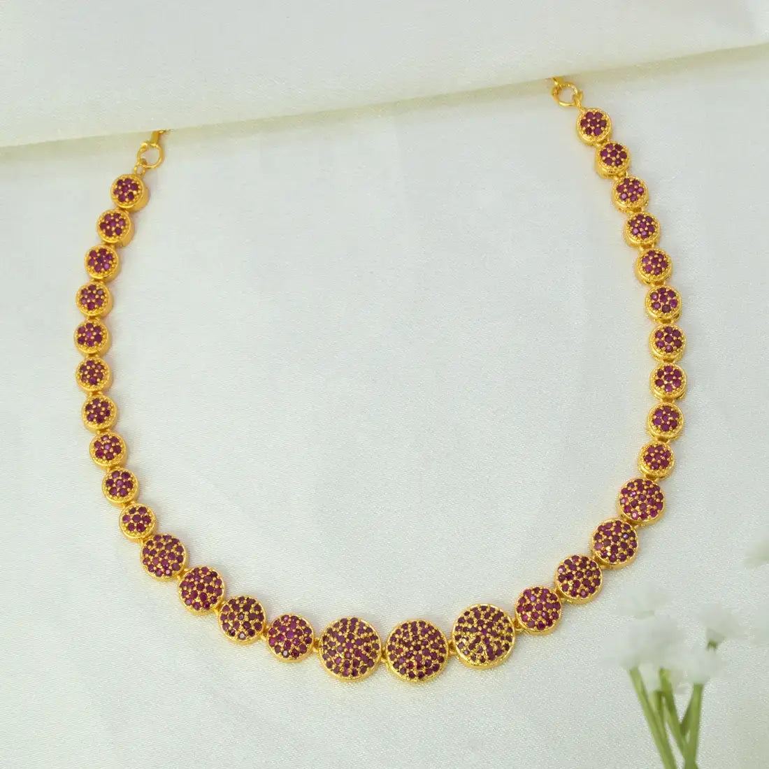 A Gold-Plated Round Design Stone Necklace featuring small ruby stones studded in clusters displayed on a soft white fabric background.