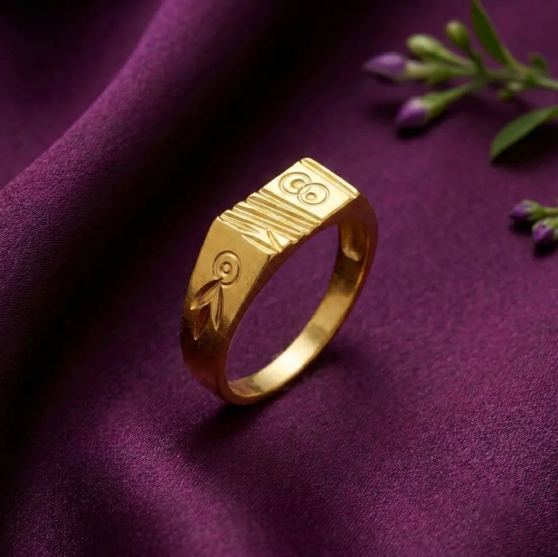 A gold ring with engraved tribal-style symbols, resting on a draped purple fabric background with small purple flowers in the corner.